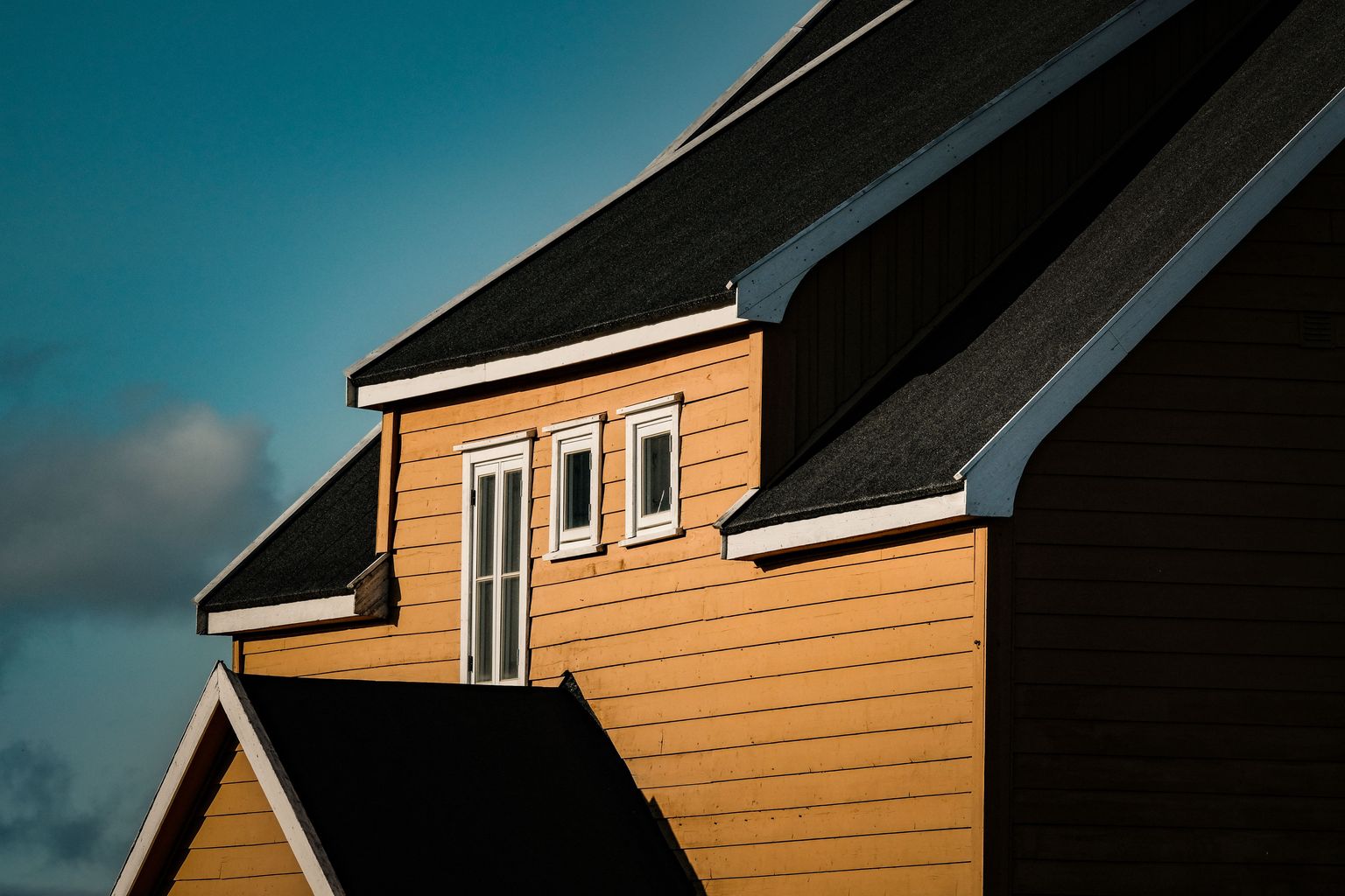 Upper floor of an orange wooden house with steep dark roofs, white trim along the roof edges and three white windows, against a blue sky